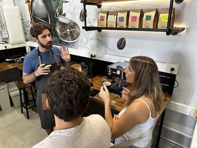 En la acogedora cafetería, el barista describió con pasión "Cata Sensorial de Café: Descubre Fragancias, Sabores y Cata Ciega" a dos clientes, enriqueciendo su apreciación con palabras sobre experiencias sensoriales en medio de ricos aromas. Cerca de allí, las bolsas de café en un estante prometían una infinidad de sabores para explorar. - ForTwo - Planes para parejas