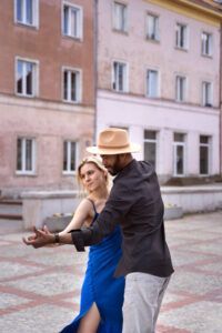 Una pareja baila elegantemente al aire libre, con la mujer con un vestido azul y el hombre con sombrero y camisa negra, capturando la esencia de Clases de Baile Latino en Monzón: Salsa Cubana contra vibrantes edificios de color rosa. - ForTwo - Planes para parejas