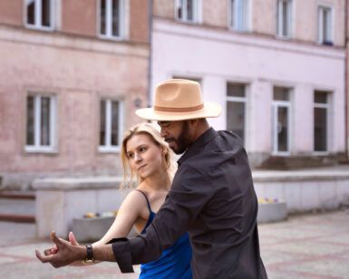 Una pareja baila elegantemente al aire libre, con la mujer con un vestido azul y el hombre con sombrero y camisa negra, capturando la esencia de Clases de Baile Latino en Monzón: Salsa Cubana contra vibrantes edificios de color rosa. - ForTwo - Planes para parejas