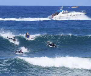 Los surfistas cabalgan hábilmente sobre las olas en el océano de Tenerife mientras un barco navega, destacando sus habilidades de surf con Clases de Surf en Tenerife para Todos los Niveles con un hermoso telón de fondo. - ForTwo - Planes para parejas