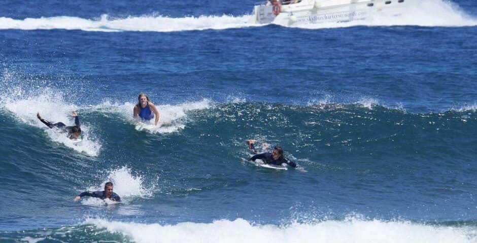 Los surfistas cabalgan hábilmente sobre las olas en el océano de Tenerife mientras un barco navega, destacando sus habilidades de surf con Clases de Surf en Tenerife para Todos los Niveles con un hermoso telón de fondo. - ForTwo - Planes para parejas
