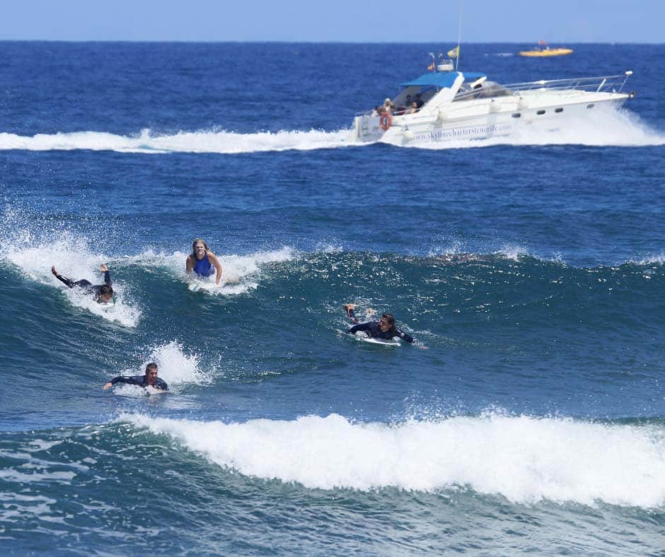 Los surfistas cabalgan hábilmente sobre las olas en el océano de Tenerife mientras un barco navega, destacando sus habilidades de surf con Clases de Surf en Tenerife para Todos los Niveles con un hermoso telón de fondo. - ForTwo - Planes para parejas