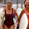 Dos mujeres sonrientes en traje de baño llevando tablas de surf caminan por un sendero de arena en Tenerife con gente al fondo, perfecto para Clases de Surf en Tenerife para Todos los Niveles. - ForTwo - Planes para parejas
