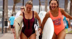 Dos mujeres sonrientes en traje de baño llevando tablas de surf caminan por un sendero de arena en Tenerife con gente al fondo, perfecto para Clases de Surf en Tenerife para Todos los Niveles. - ForTwo - Planes para parejas