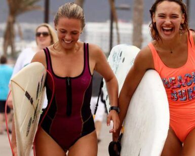 Dos mujeres sonrientes en traje de baño llevando tablas de surf caminan por un sendero de arena en Tenerife con gente al fondo, perfecto para Clases de Surf en Tenerife para Todos los Niveles. - ForTwo - Planes para parejas