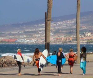 Cinco surfistas con tablas caminan por un sendero costero en Tenerife, rodeados de palmeras y edificios distantes. Olas perfectas los esperan, un indicio de las emocionantes "Clases de Surf en Tenerife para Todos los Niveles". - ForTwo - Planes para parejas