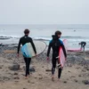 Dos surfistas, sosteniendo sus tablas, cruzan la playa rocosa de Las Américas en Tenerife, ansiosos por abrazar las olas y perfeccionar sus habilidades después de una vigorizante mañana de "Clases de Surf para parejas: Las Américas, Tenerife. - ForTwo - Planes para parejas