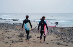 Dos surfistas, sosteniendo sus tablas, cruzan la playa rocosa de Las Américas en Tenerife, ansiosos por abrazar las olas y perfeccionar sus habilidades después de una vigorizante mañana de "Clases de Surf para parejas: Las Américas, Tenerife. - ForTwo - Planes para parejas