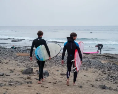 Dos surfistas, sosteniendo sus tablas, cruzan la playa rocosa de Las Américas en Tenerife, ansiosos por abrazar las olas y perfeccionar sus habilidades después de una vigorizante mañana de "Clases de Surf para parejas: Las Américas, Tenerife. - ForTwo - Planes para parejas