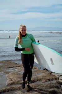 Un surfista sonríe con una camiseta verde y un traje de neopreno negro, sosteniendo su tabla mientras disfruta de las vistas de la playa rocosa de Tenerife durante las "Clases de Surf para parejas: Las Américas, Tenerife. - ForTwo - Planes para parejas