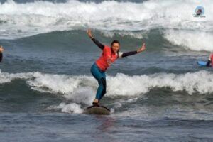 Una persona monta pequeñas olas con los brazos extendidos, vistiendo una camisa roja y pantalones azules, disfrutando de la emoción en Clases de surf privadas en Volcano Surf School Tenerife. - ForTwo - Planes para parejas