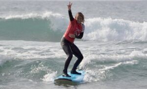 Un surfista con camiseta roja surfea una ola cerca de Tenerife, sonriendo y haciendo el símbolo de la paz. Es el día perfecto para las clases de surf privadas en Volcano Surf School Tenerife. - ForTwo - Planes para parejas