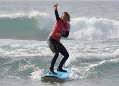 Un surfista con camiseta roja surfea una ola cerca de Tenerife, sonriendo y haciendo el símbolo de la paz. Es el día perfecto para las clases de surf privadas en Volcano Surf School Tenerife. - ForTwo - Planes para parejas
