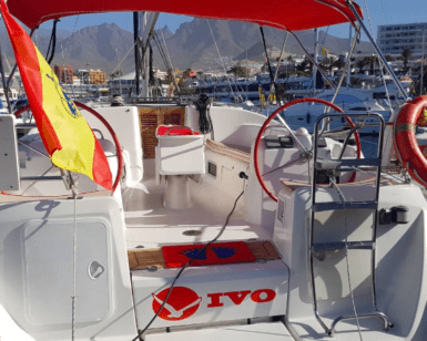 Un impresionante velero con toldo rojo y ruedas de timón dobles está atracado en el vibrante puerto deportivo de Costa Adeje, adornado con una bandera española en la popa, listo para su próximo "Crucero de 3 Horas en Costa Adeje". - ForTwo - Planes para parejas