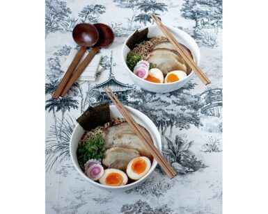 Dos tazones de ramen con huevos, carne y algas descansan sobre una superficie blanca y negra, flanqueados por palillos de madera, creando una fusión de sabores que evocan la experiencia del "Curso: Domina la Olla Rápida y Cocina Sabores del Mundo". - ForTwo - Planes para parejas