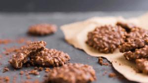 Un primer plano de galletas de chocolate con nueces sobre papel vegetal en la cocina. Una galleta está parcialmente consumida, y hay migas esparcidas sobre un fondo que destaca "Curso de Cocina: Domina Carnes, Pescados y Arroces". - ForTwo - Planes para parejas