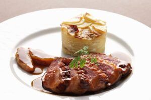El pato asado en rodajas con salsa, servido elegantemente junto con un gratinado de patatas en capas y guarnición de perejil, parece un plato del "Curso de Cocina Francesa: Descubre el Arte Culinario con Mélanie Dyé". - ForTwo - Planes para parejas