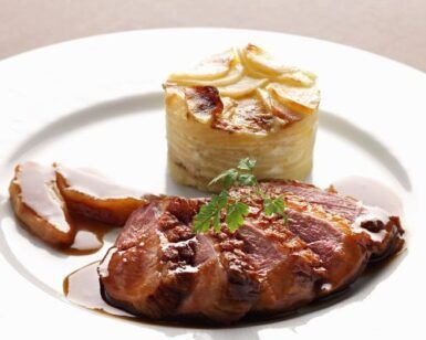El pato asado en rodajas con salsa, servido elegantemente junto con un gratinado de patatas en capas y guarnición de perejil, parece un plato del "Curso de Cocina Francesa: Descubre el Arte Culinario con Mélanie Dyé". - ForTwo - Planes para parejas