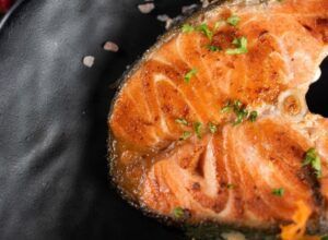 El "Curso de cocina: Postureo Gastronómico" presenta un filete de salmón a la parrilla en un plato negro, adornado con perejil y cristales de sal: una obra maestra culinaria para los amantes de la alta cocina. - ForTwo - Planes para parejas