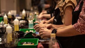Los participantes, ataviados con delantales, están ocupados preparando comida en el Curso de cocina: Postureo Gastronómico, con ingredientes frescos y utensilios cuidadosamente dispuestos en el mostrador. - ForTwo - Planes para parejas
