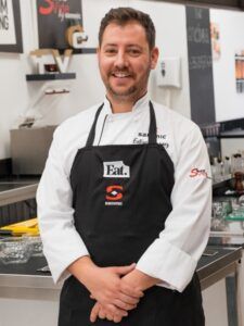 Un chef sonriente con un delantal negro se encuentra de pie con confianza y los brazos cruzados en una cocina moderna, mostrando su maestría en el "Curso de cocina: Postureo Gastronómico". - ForTwo - Planes para parejas