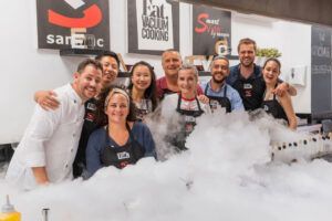 Un grupo de personas en una cocina, sonriendo entre el humo y con delantales que dicen Eat Vacuum Cooking, claramente disfrutando de su aventura gastronómica del Curso de cocina: Postureo Gastronómico. - ForTwo - Planes para parejas