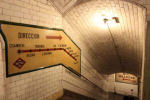 Las paredes de azulejos de la poco iluminada Estación Fantasma de Chamberí de Madrid muestran carteles antiguos del metro con nombres de estaciones y flechas, que capturan el encanto de los Años 20. Titulado "Descubre la Estación Fantasma de Chamberí: Un Viaje al Madrid de los Años 20". - ForTwo - Planes para parejas