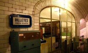 Una antigua taquilla de metro con un cartel de "Billetes Precio Exacto", rodeada de paredes de azulejos y cristaleras, evoca el encanto de la Estación Fantasma de Chamberí de Madrid de los años 20. Descubre este viaje atemporal a través de nuestro producto: Descubre la Estación Fantasma de Chamberí: Un Viaje al Madrid de los Años 20. - ForTwo - Planes para parejas