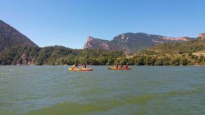 Los participantes de la "Excursión Guiada en Kayak en Coll de Nargó: Naturaleza y Aventura en el Pantano" se deslizan por las serenas aguas, rodeados de frondosos bosques y majestuosas montañas bajo un claro cielo azul. - ForTwo - Planes para parejas