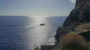 Bajo un cielo despejado, un barco navega por el océano iluminado por el sol cerca de un acantilado rocoso, proporcionando un lugar ideal para el avistamiento de ballenas en Canarias, mostrando la majestuosa belleza de las ballenas de la región. - ForTwo - Planes para parejas