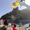 La gente en un barco cerca de una isla rocosa en Canarias, con una bandera amarilla ondeando arriba, espera ansiosamente un "Avistamiento de ballenas en Canarias". - ForTwo - Planes para parejas