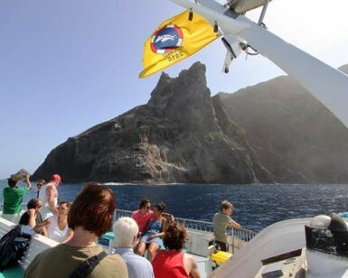 La gente en un barco cerca de una isla rocosa en Canarias, con una bandera amarilla ondeando arriba, espera ansiosamente un "Avistamiento de ballenas en Canarias". - ForTwo - Planes para parejas