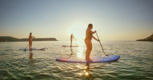 Tres personas practican paddle surf con gracia en las tranquilas aguas de Ibiza al atardecer, recortadas por el sol durante "Excursiones de SUP en Ibiza: Atardecer sobre el agua. - ForTwo - Planes para parejas
