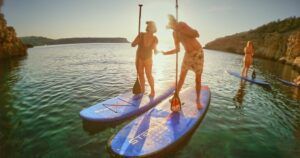 Dos personas disfrutan del paddleboarding al atardecer en un mar tranquilo en Ibiza, rodeados de pintorescos acantilados y otro entusiasta del SUP, como parte de las "Excursiones de SUP en Ibiza: Atardecer sobre el agua. - ForTwo - Planes para parejas