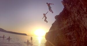 Mientras el sol se pone en Ibiza, dos aventureros saltan al mar desde el acantilado mientras los paddleboarders cercanos de "Excursiones de SUP en Ibiza: Atardecer sobre el agua" flotan y observan cómo se desarrolla la escena. - ForTwo - Planes para parejas