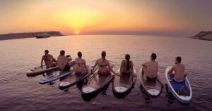 Siete personas sentadas en tablas de paddle surf durante las "Excursiones de SUP en Ibiza: Atardecer sobre el agua", cautivadas por la puesta de sol. El mar en calma, las colinas lejanas y el yate crean un entorno inolvidable y sereno. - ForTwo - Planes para parejas