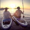 Dos personas practicando paddle surf al atardecer, con veleros y una costa espectacular. Esta aventura de SUP encarna la magia de "Excursiones de SUP en Ibiza: Atardecer sobre el agua", ideal para quienes sueñan con explorar Ibiza. - ForTwo - Planes para parejas