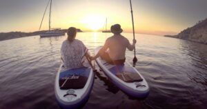 Dos personas practicando paddle surf al atardecer, con veleros y una costa espectacular. Esta aventura de SUP encarna la magia de "Excursiones de SUP en Ibiza: Atardecer sobre el agua", ideal para quienes sueñan con explorar Ibiza. - ForTwo - Planes para parejas