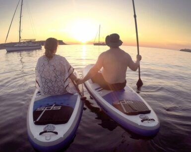 Dos personas practicando paddle surf al atardecer, con veleros y una costa espectacular. Esta aventura de SUP encarna la magia de "Excursiones de SUP en Ibiza: Atardecer sobre el agua", ideal para quienes sueñan con explorar Ibiza. - ForTwo - Planes para parejas
