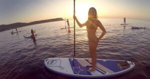 Una mujer flota tranquilamente en su tabla de paddle surf al atardecer, recortada contra el mar tranquilo mientras otros se deslizan a su lado, capturando la magia de "Excursiones de SUP en Ibiza: Atardecer sobre el agua. - ForTwo - Planes para parejas