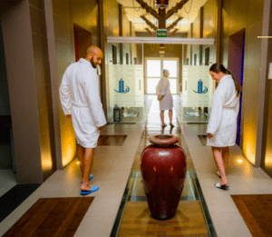 Dos personas en batas disfrutan de la máxima relajación paseando por el pasillo de un spa, donde la Experiencia de Relax en Pareja crea un ambiente tranquilo con un gran jarrón central. - ForTwo - Planes para parejas