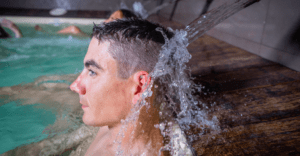 Alguien disfruta de la "Experiencia de Relax en Pareja", relajándose bajo el agua en cascada de una piscina desde una tranquila vista lateral. - ForTwo - Planes para parejas