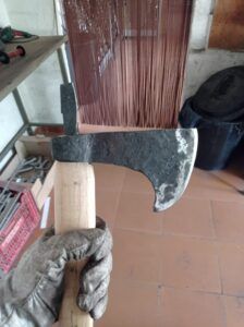 En un taller, una mano enguantada sostiene un hacha de metal con mango de madera, invitándote a "Forja Tu Propia Hacha: Una Experiencia Única". - ForTwo - Planes para parejas