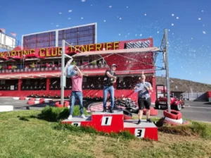 Tres hombres celebran en un podio en Karting en Tenerife Sur (Los Cristianos), rociando champán con la pista y el edificio del club de fondo. - ForTwo - Planes para parejas