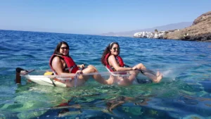 Dos personas con chalecos salvavidas rojos se sientan en un Kayak Transparente en Radazul: Explora el fondo marino de Tenerife, deslizándose sobre las soleadas aguas azules cerca de la pintoresca costa de Radazul, revelando la fascinante vida marina debajo. - ForTwo - Planes para parejas