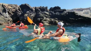 Los kayakistas en las cristalinas aguas de Radazul, con chalecos salvavidas y remando con remos naranjas, disfrutan de un impresionante telón de fondo mientras exploran el fascinante fondo marino de Tenerife con el Kayak Transparente en Radazul. - ForTwo - Planes para parejas