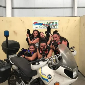 Cinco mujeres adoptan poses dinámicas con pistolas láser detrás de una motocicleta BMW, encarnando la esencia de "Láser Combat: Vive la emoción del combate en Gijón" en un estadio cubierto, mostrando el emocionante espíritu del combate. - ForTwo - Planes para parejas