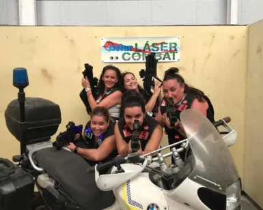 Cinco mujeres adoptan poses dinámicas con pistolas láser detrás de una motocicleta BMW, encarnando la esencia de "Láser Combat: Vive la emoción del combate en Gijón" en un estadio cubierto, mostrando el emocionante espíritu del combate. - ForTwo - Planes para parejas