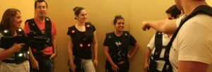 Un grupo de personas con chalecos de laser tag se reúne, y una persona hace un gesto hacia ellos, listos para una emocionante ronda de Láser Combat: Vive la emoción del combate en Gijón. - ForTwo - Planes para parejas