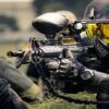 En medio de una intensa partida de Mallorca Paintball, un jugador equipado de forma completa apunta su arma con precisión, mientras sus compañeros elaboran estrategias con el telón de fondo de los 5 Campos Tematizados. - ForTwo - Planes para parejas
